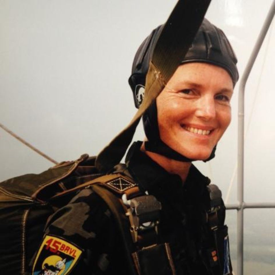 A smiling person wearing a helmet and parachute gear stands outdoors. They are dressed in a uniform with various patches, including one with a cartoon character and "15 BAYL" written on it. The sky is visible in the background.