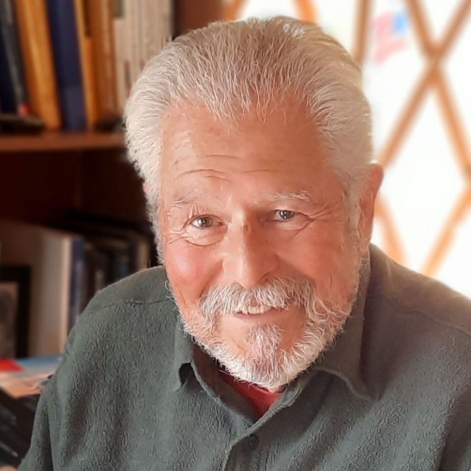 Elderly man with white hair and beard smiles warmly, sitting indoors. Books are visible in the background, suggesting a cozy, studious environment. The man is wearing a dark green sweater.