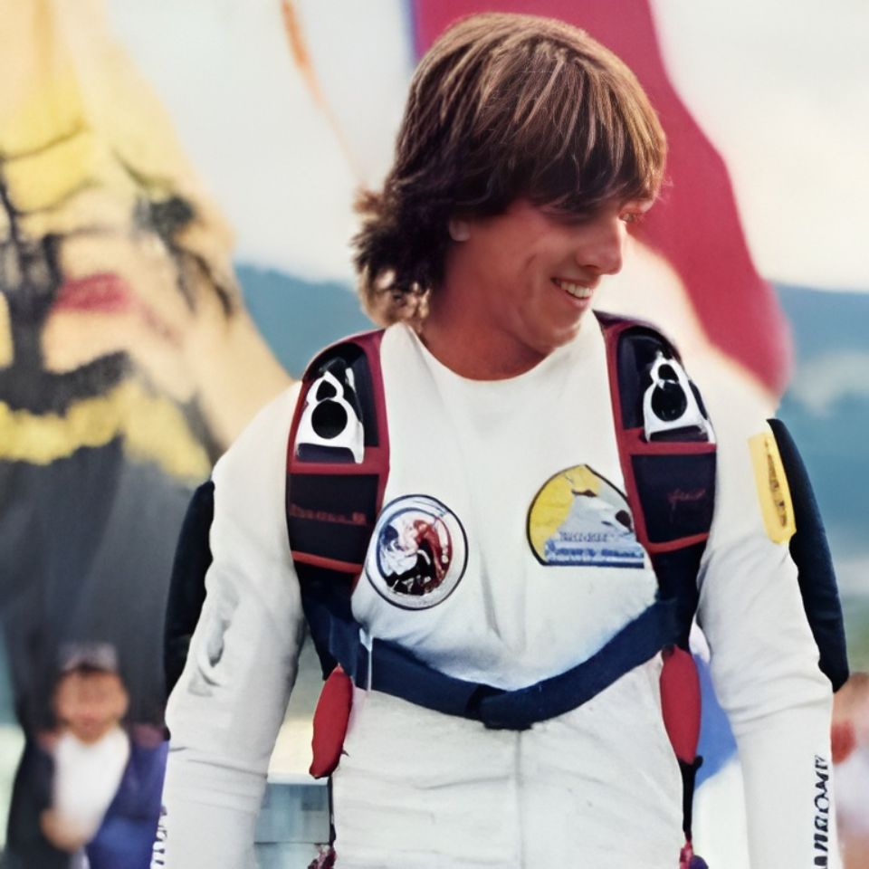 A man with long hair is smiling, wearing a white shirt with various patches and a black harness. In the background, there are flags and a blurred figure. The setting appears to be outdoors.