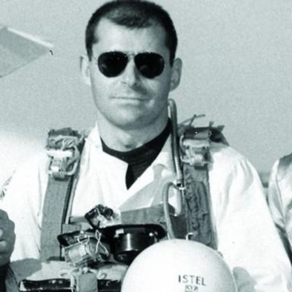 A man wearing a flight suit and sunglasses holds a helmet with "ISTEL" written on it. The image is black and white, and he appears to be standing outdoors.