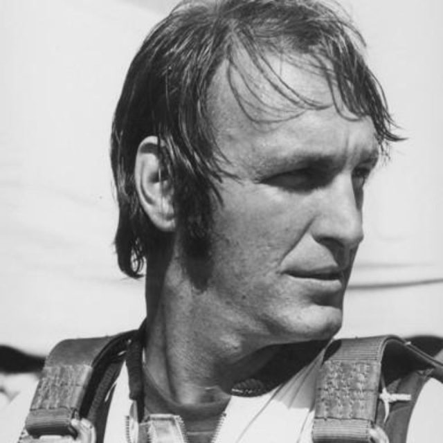 A black-and-white image of a man with short hair looking to the right. He's wearing a parachute harness over a shirt. The background is blurred, emphasizing his profile and focused expression.