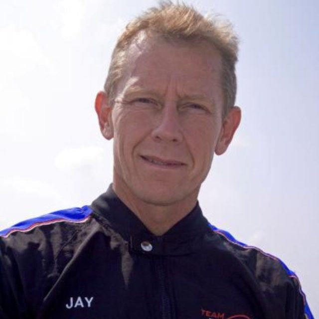 A person wearing a racing suit with "Jay" embroidered on it stands outdoors against a clear sky. The suit features blue and red accents. The person has short hair and appears to be looking into the camera.