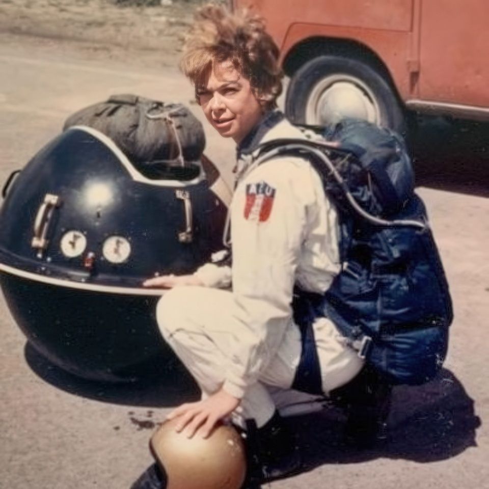 A person in a white jumpsuit and backpack crouches beside a spherical device with dials and gauges. A helmet rests on the ground nearby. A red vehicle is in the background.