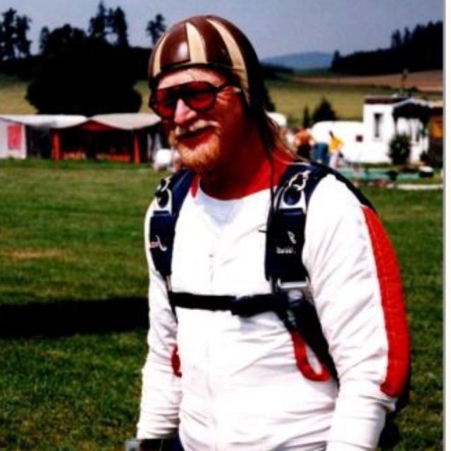 A person wearing a skydiving suit and helmet stands smiling on a grassy field. The suit is white with red accents, and they are wearing sunglasses. In the background, there are trees and a few small buildings.