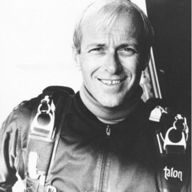 A smiling person wearing a parachute harness and jumpsuit stands in front of a plain background. The harness is marked with the word "talon.