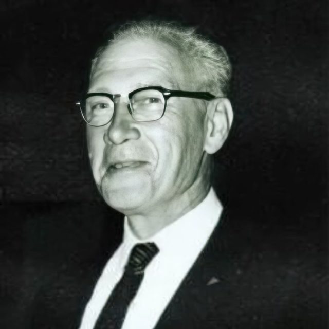 An older man wearing glasses, a suit, and a tie, smiles while facing the camera. The background is dark, highlighting the man's face and attire.