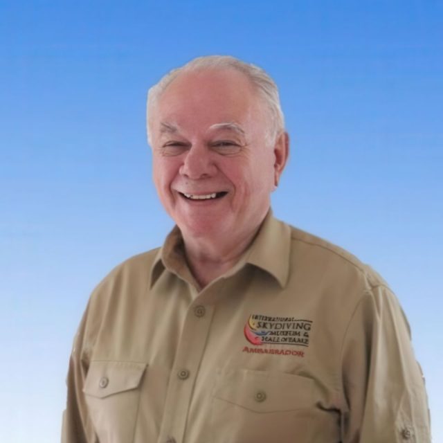 An elderly man with white hair is smiling warmly against a gradient blue background. He is wearing a beige button-up shirt with embroidered text and logos related to skydiving.