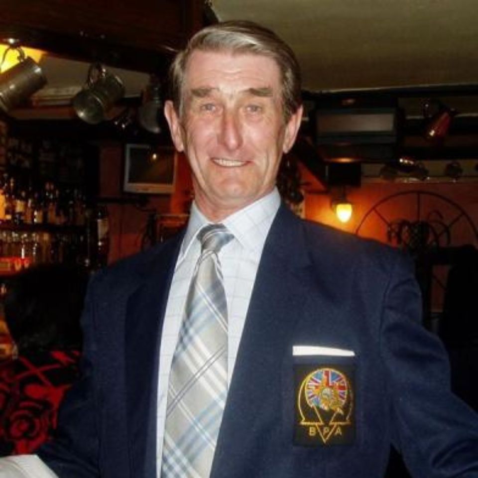 A smiling man in a plaid shirt and tie, wearing a navy blazer with a crest on the pocket, stands in a cozy bar or pub setting. The background shows dim lighting and various bottles.