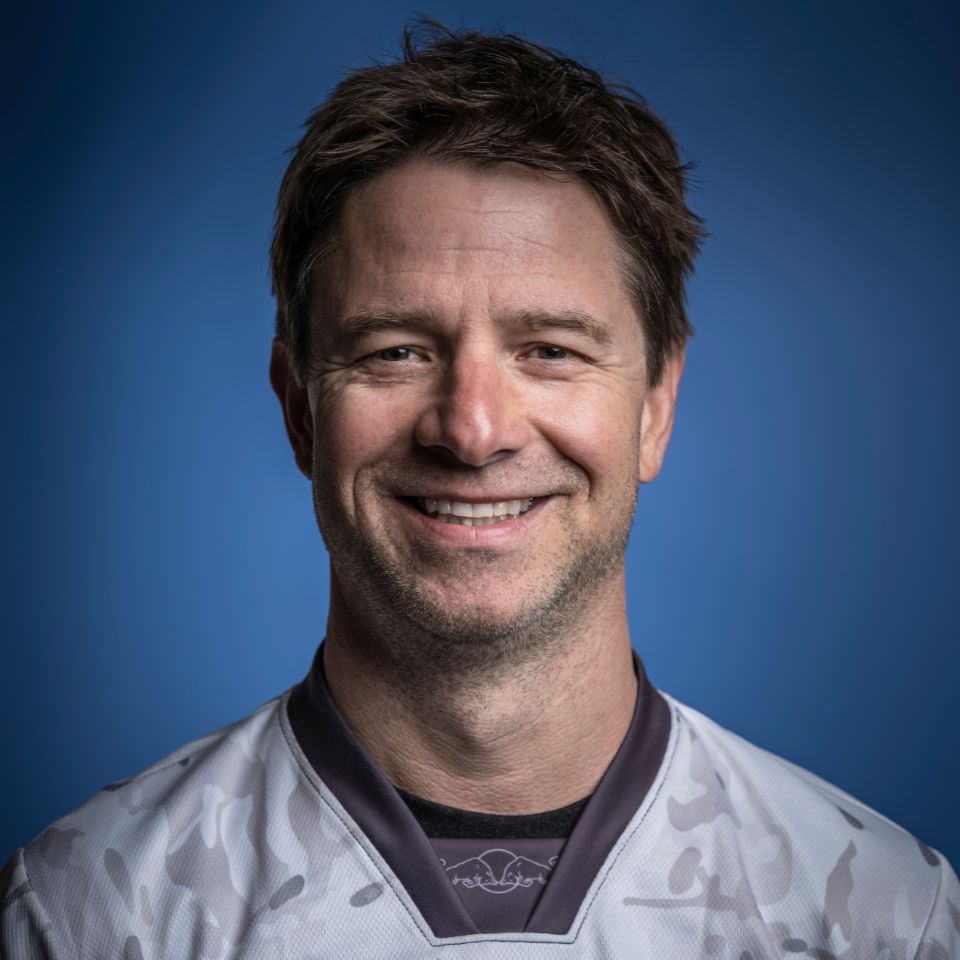 A person with short brown hair smiles warmly at the camera. They are wearing a gray and white patterned shirt against a blue background.