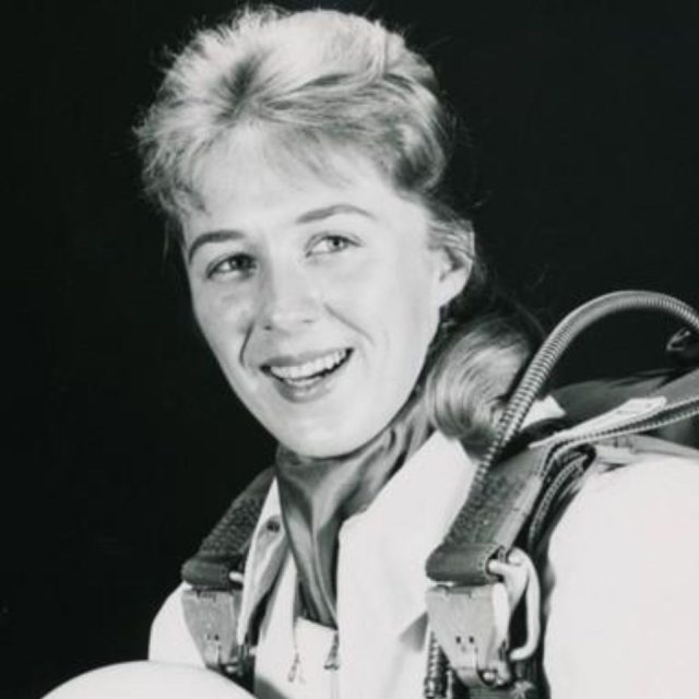 A black and white photo of a smiling woman in a spacesuit. She has light hair pulled back and is holding a helmet. The suit features various straps and connectors. The background is plain and dark, highlighting her expression and attire.