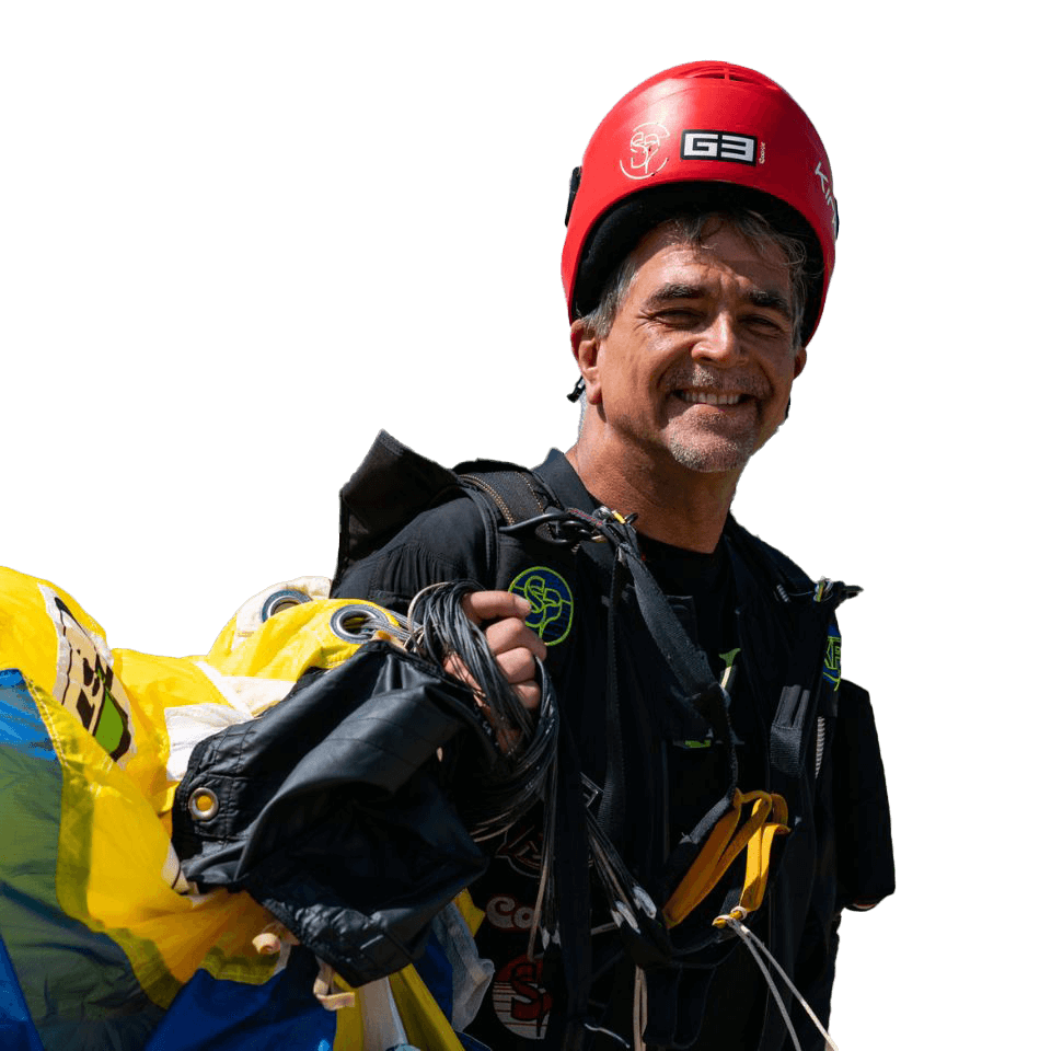 A person wearing a red helmet and black jumpsuit smiles while holding a partially folded parachute.