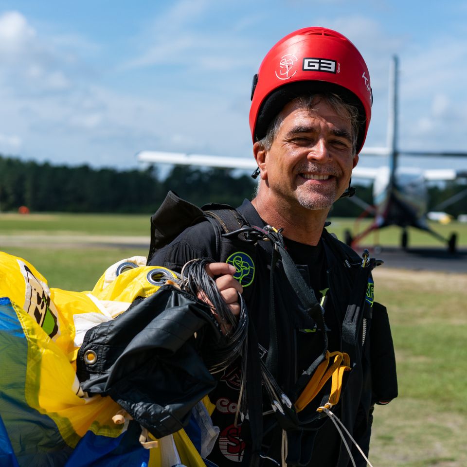 A person in skydiving gear smiles while holding a parachute, standing on a grass field. They wear a red helmet, and a plane is visible in the background. The sky is clear and sunny.