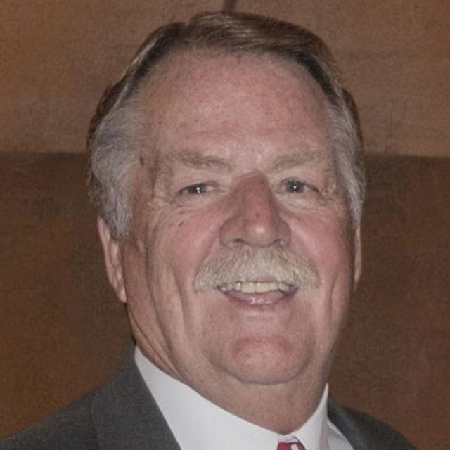 Smiling older man with gray hair and a mustache, wearing a suit and tie, against a brown background.