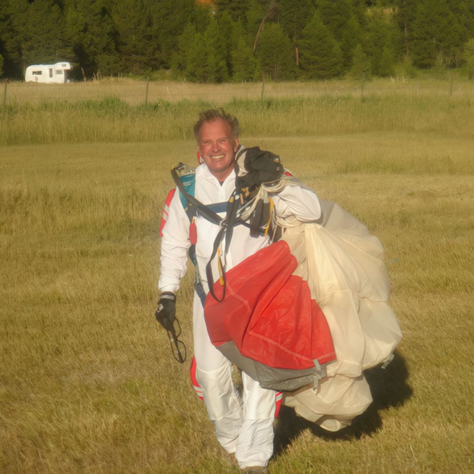 A person wearing a white and red jumpsuit smiles while carrying a parachute across a grassy field. Trees and a white trailer are visible in the background under clear skies.