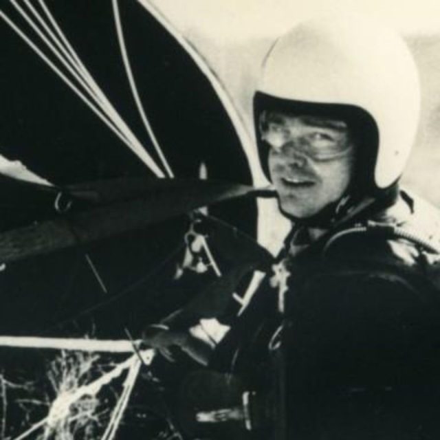 A person wearing a helmet and glasses is harnessed to a hang glider, preparing for flight. The black and white image captures an adventurous moment with focused expression and parachute cables visible.