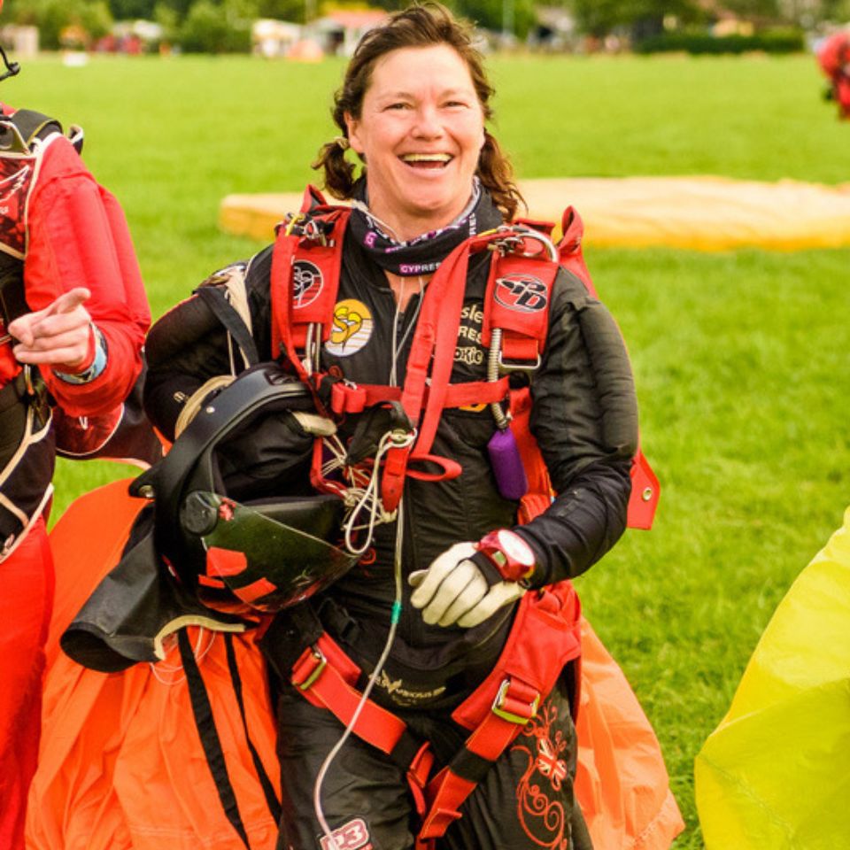 A smiling person in a parachuting outfit, holding a helmet, stands on a grassy field. They are wearing bright red and black gear, with a parachute visible in the background. Two other people are partially visible beside them.