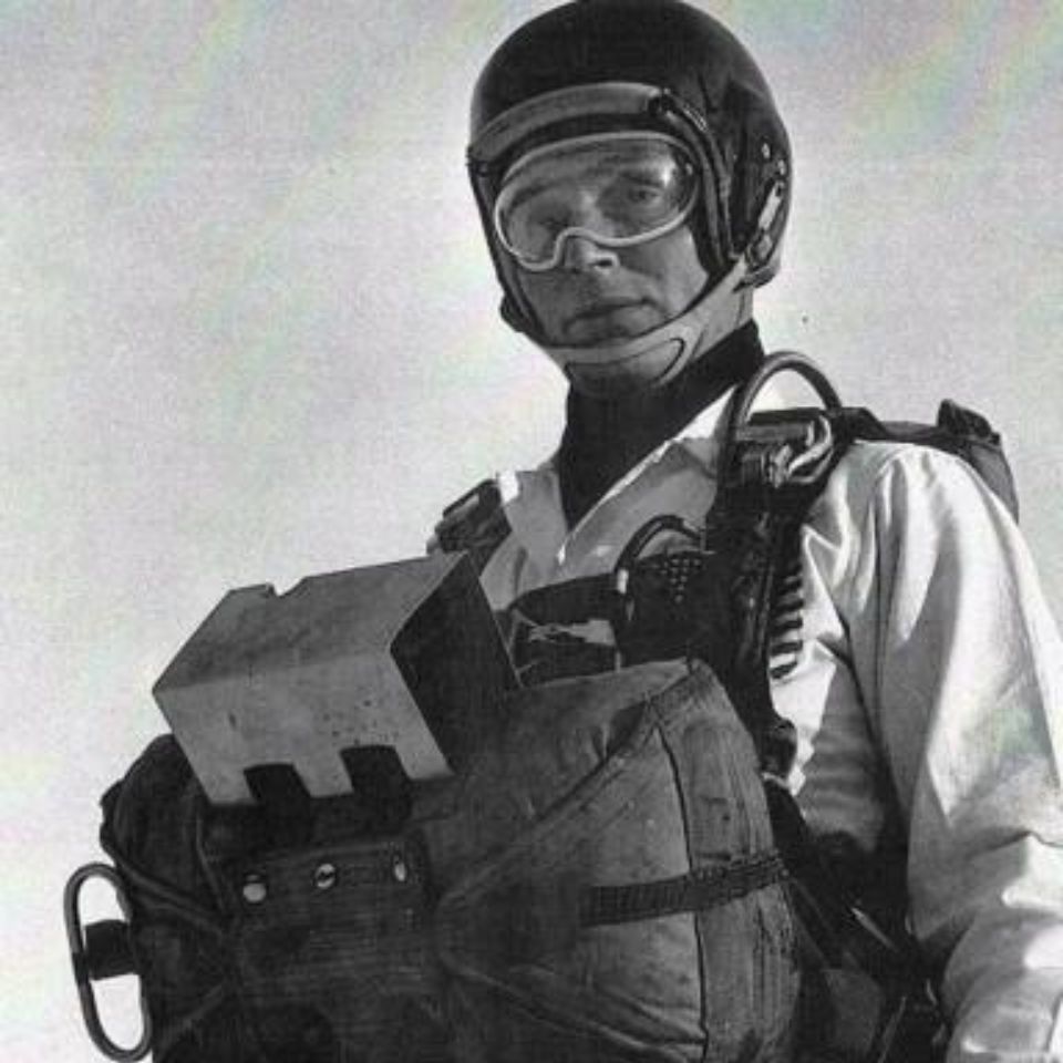 A person wearing a helmet and goggles stands with a parachute strapped to their chest. The black and white image shows them looking towards the camera, wearing a jacket and protective gear against a plain sky background.