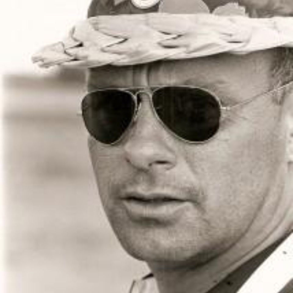 A person wearing aviator sunglasses and a woven hat gazes into the distance. The image is in black and white, providing a classic and timeless feel. The background is blurred, focusing attention on the individual's facial expression.