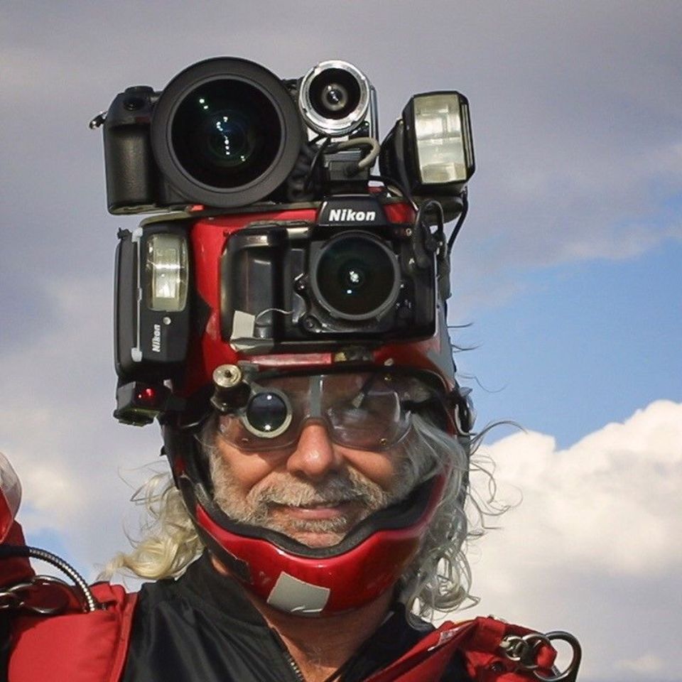 A person with gray hair wearing a helmet creatively rigged with multiple cameras and flashes. They have glasses and are set against a partly cloudy sky.