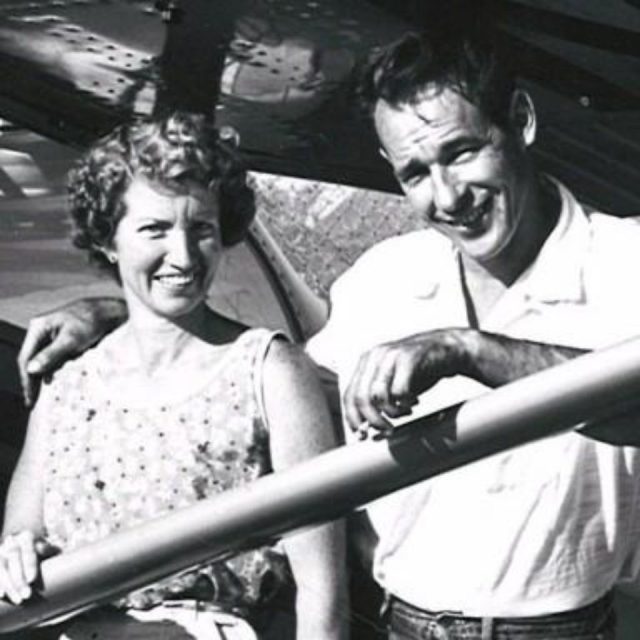 A black and white photo of a smiling couple. The man, in a short-sleeved shirt, leans on a railing with his arm around the woman's shoulders. The woman, wearing a sleeveless polka dot dress, stands beside him. They are outdoors, possibly on a boat.