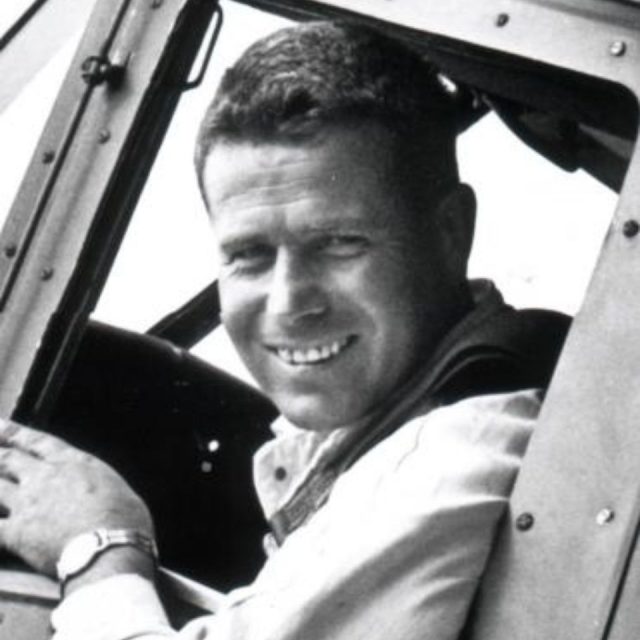 A man smiling while sitting inside a vehicle, appearing to be a cockpit or cabin. He is wearing a light-colored shirt and a watch on his left wrist. The image is black and white.