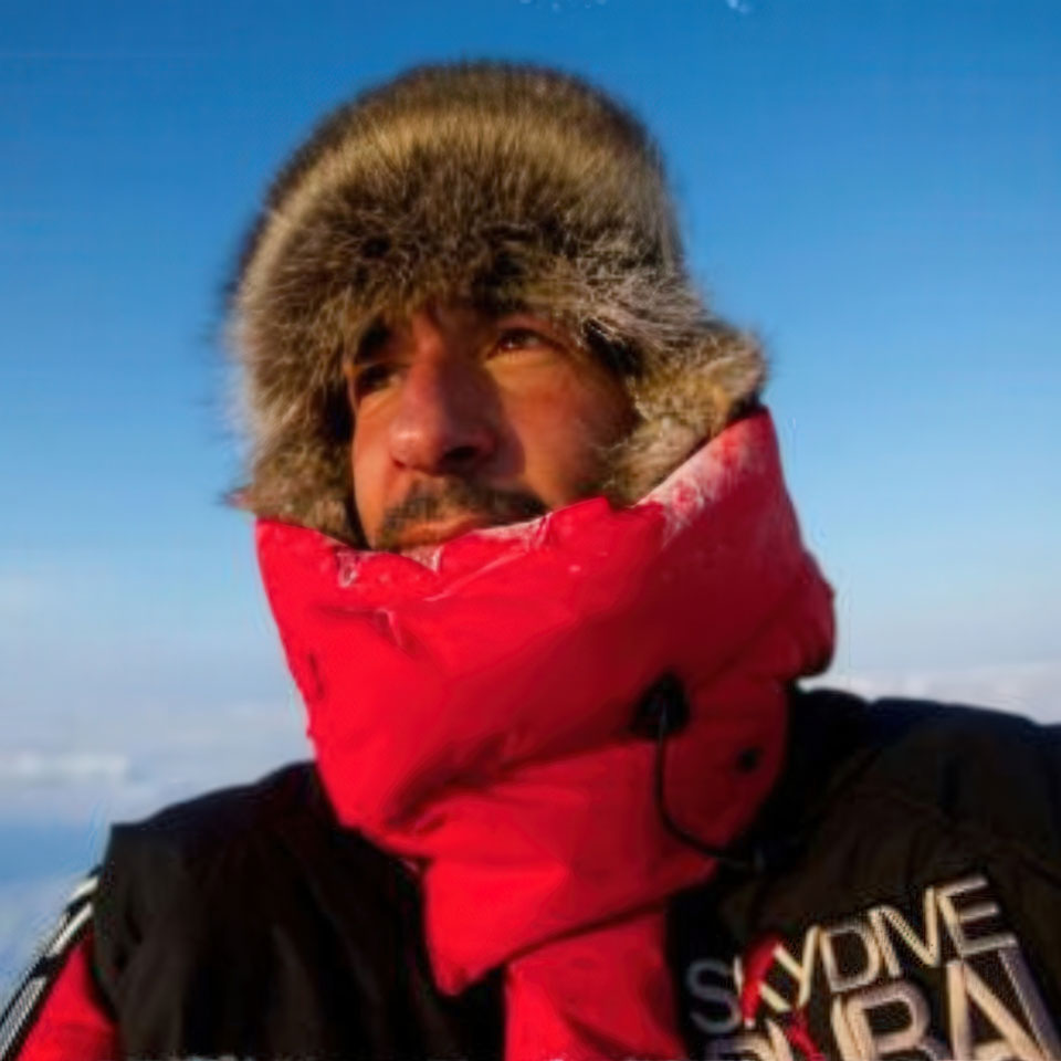 A person dressed in a red jacket and furry hat stands against a clear blue sky. The jacket has the text "Skydive Polar" on it. The person looks into the distance, with a calm expression on their face.