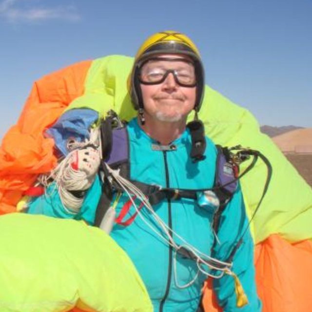 A person wearing a jumpsuit, helmet, and goggles smiles while holding a bright neon parachute. The background features a clear blue sky and desert terrain.