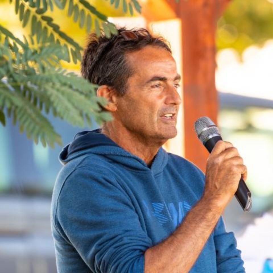 A man in a blue hoodie holds a microphone while speaking outdoors. He's standing under a tree, and there is soft sunlight and greenery in the background.