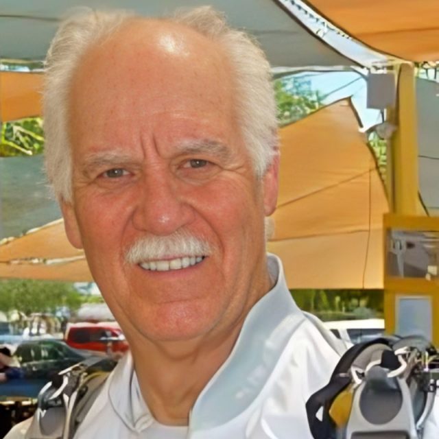 An elderly man with white hair and a mustache smiles warmly. He's wearing a light-colored jacket and has a parachute harness on his shoulders. The background shows a sunny outdoor setting with cars and shade sails.