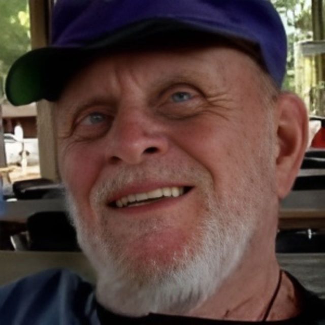 A bearded man wearing a blue cap smiles warmly while sitting indoors. The background is softly blurred, with hints of outdoor scenery.