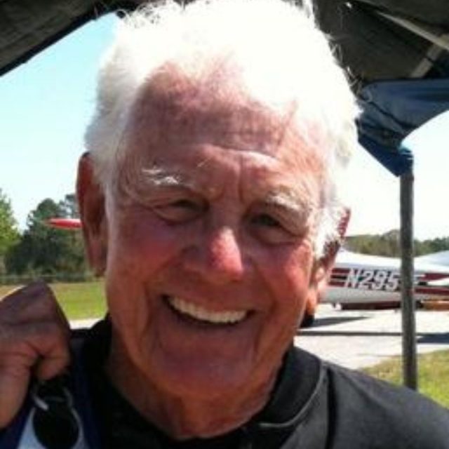 An older man with white hair smiles outdoors in front of an airplane. He wears a dark shirt and carries a jacket over his shoulder. The background includes grassy areas and part of a small aircraft.