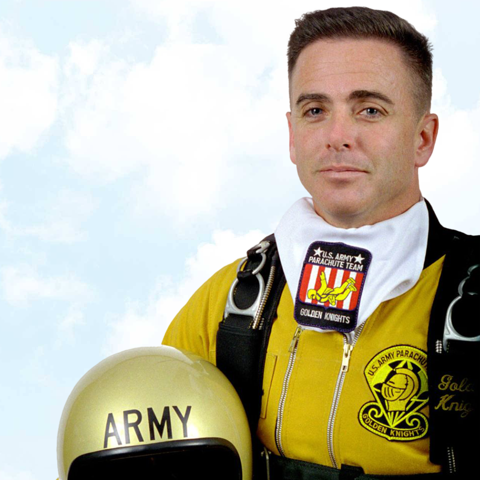 A person in a yellow jumpsuit with patches for the U.S. Army Parachute Team and Golden Knights stands holding a gold helmet labeled "Army." The person is against a backdrop of a clear blue sky with clouds.