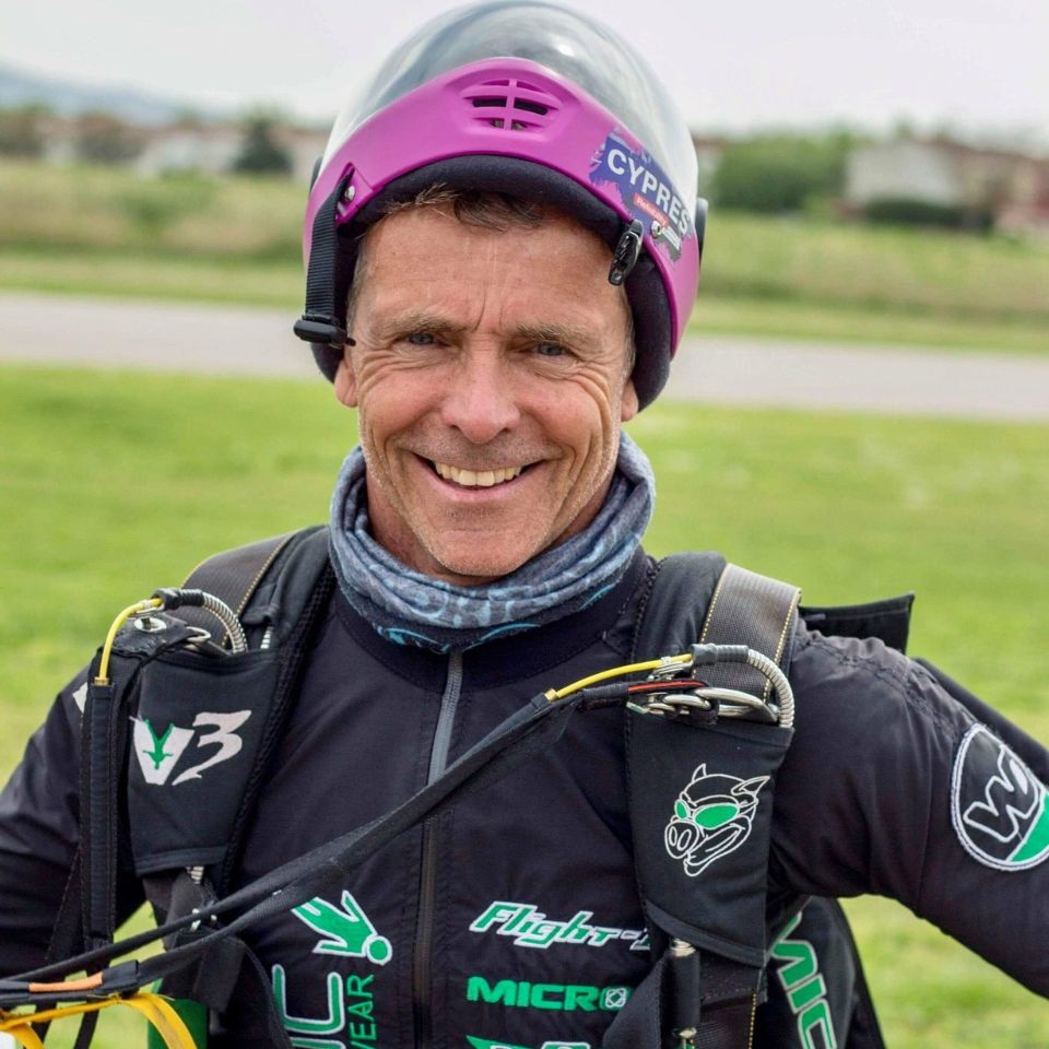 A person wearing a skydiving jumpsuit and helmet stands outdoors. The helmet is purple with a clear visor. The jumpsuit is black with various logos. The person is smiling, and the background is grassy with an out-of-focus horizon.