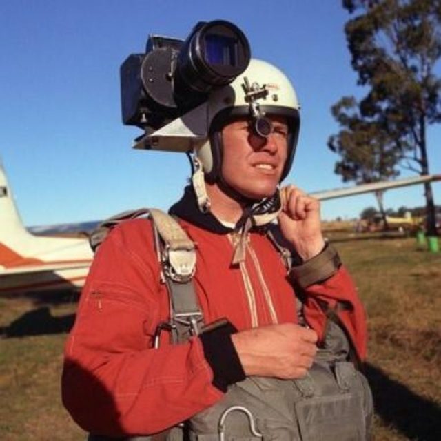 A person wearing a white helmet with a large camera mounted on it stands outdoors in front of a small plane, wearing a red jacket and parachute harness. They are looking into the distance under a clear blue sky.