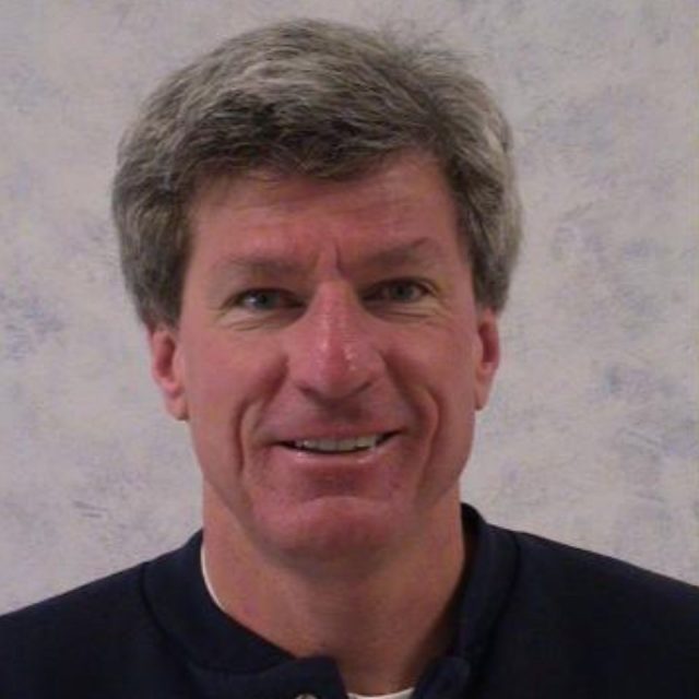 Portrait of a middle-aged man with short gray hair, wearing a dark top, smiling at the camera against a light-colored, textured background.