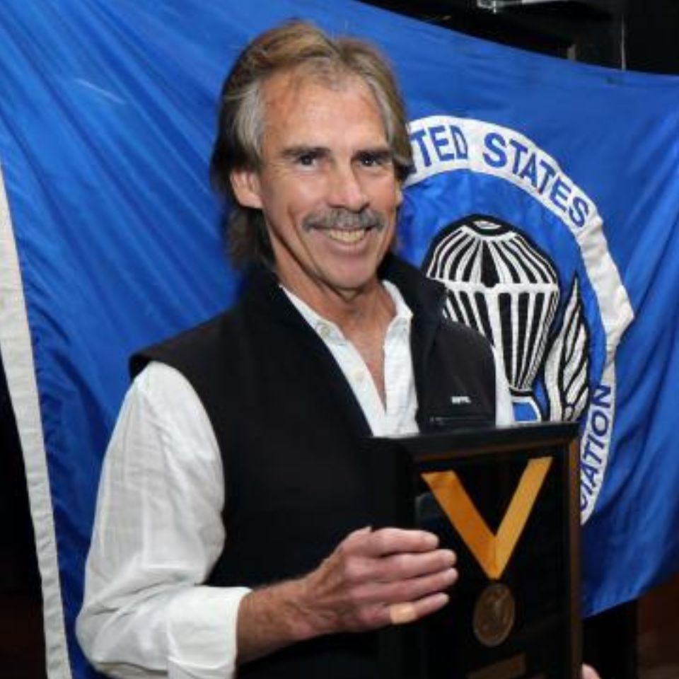 A smiling man with a mustache holds a framed award, standing in front of a blue United States flag with a military insignia. He wears a white shirt and dark vest.