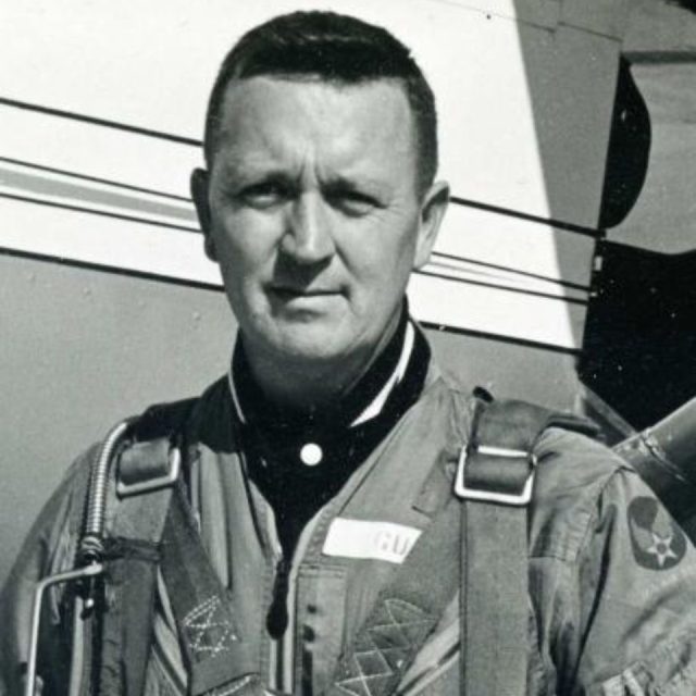 A black and white photo of a man in a military flight suit with harnesses, standing in front of an aircraft. He has short hair, and the background shows part of the plane's body with a visible star emblem on his uniform.