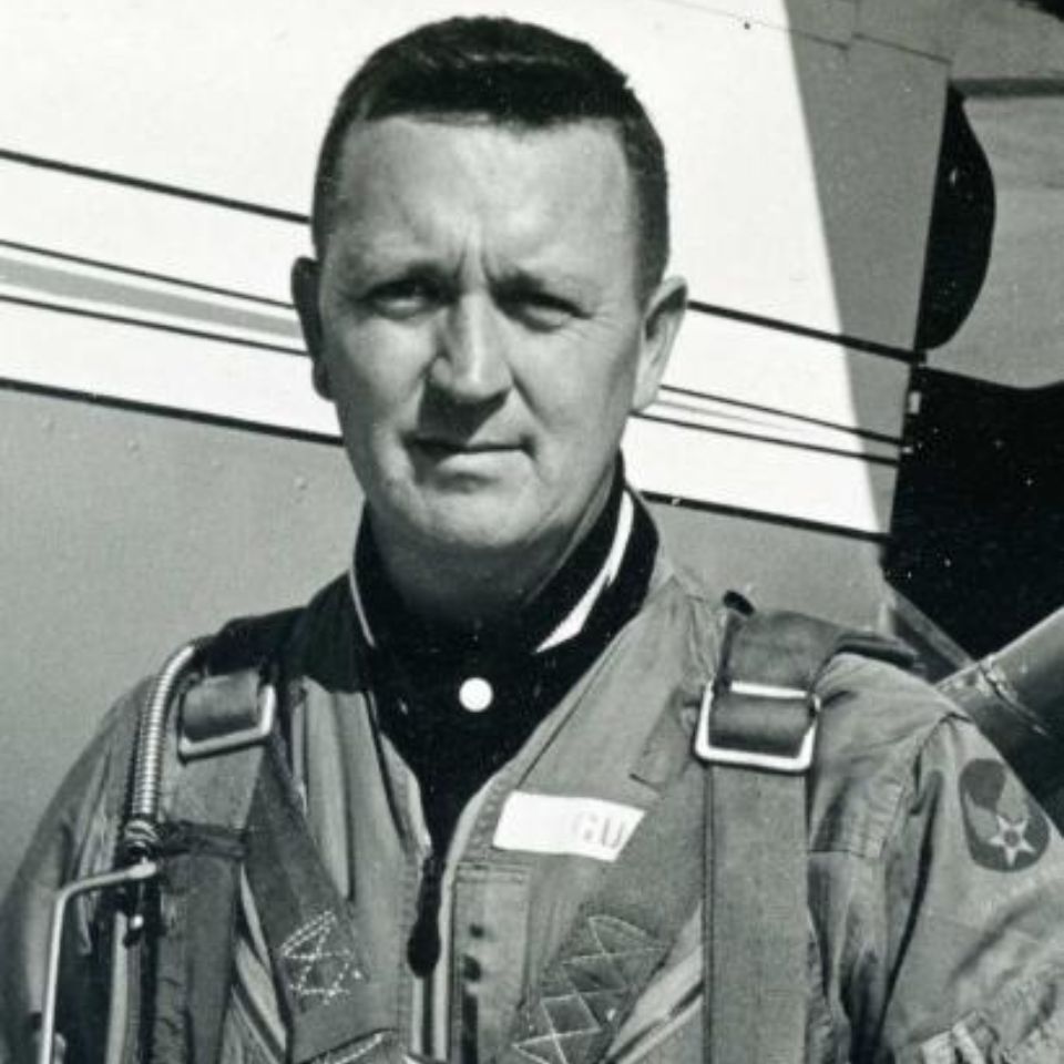 A black and white photo of a man in a military flight suit with harnesses, standing in front of an aircraft. He has short hair, and the background shows part of the plane's body with a visible star emblem on his uniform.