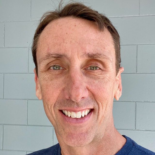 A man with short brown hair smiles at the camera. He is wearing a dark blue shirt and stands against a light blue brick wall.