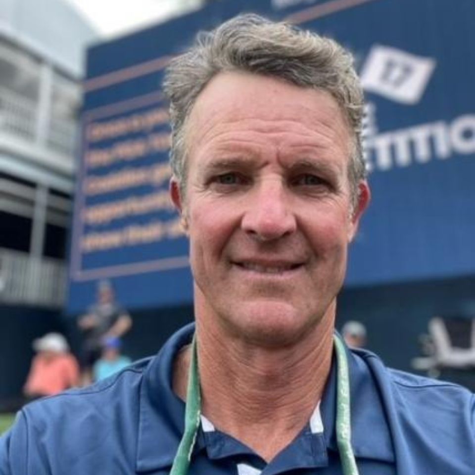 A man with short, gray hair is smiling at the camera. He is wearing a blue collared shirt and has a green lanyard around his neck. The background shows a blurred outdoor area with a billboard displaying some text.