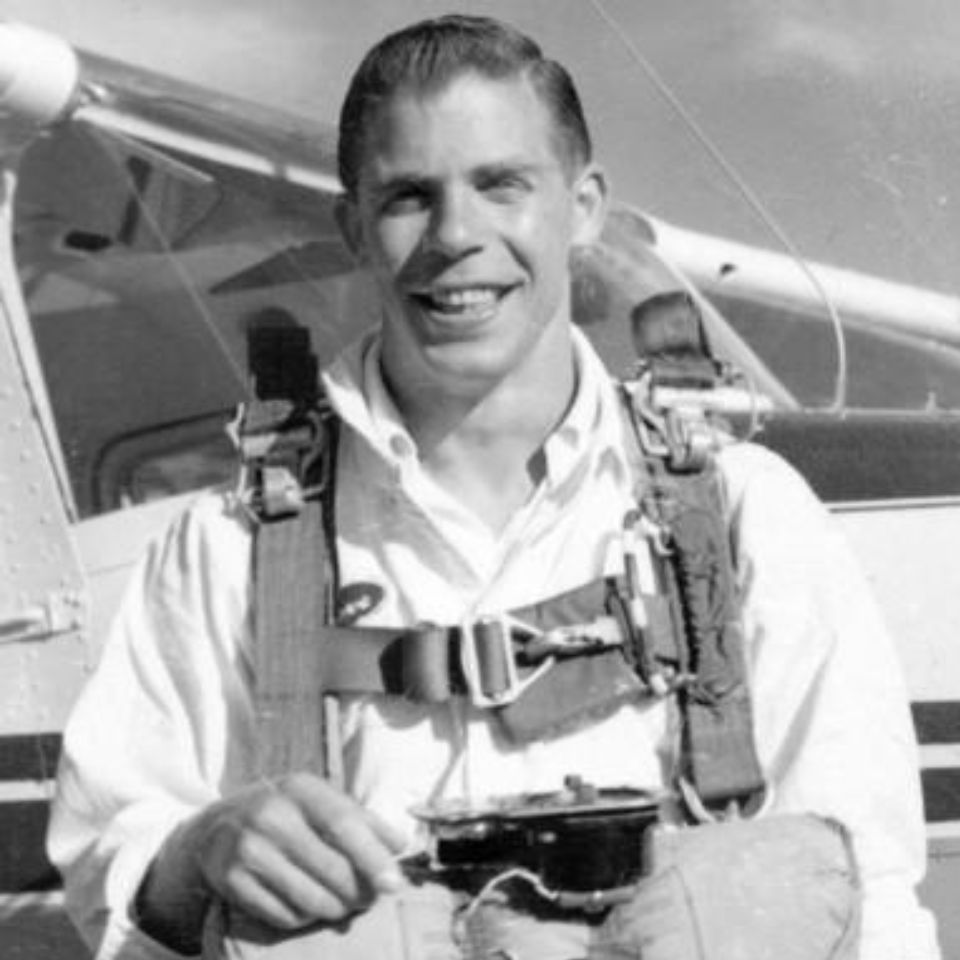 A smiling person in a parachute harness stands in front of a small aircraft. The image is in black and white, suggesting a vintage or historical context. The individual is holding a piece of equipment, possibly related to parachuting.