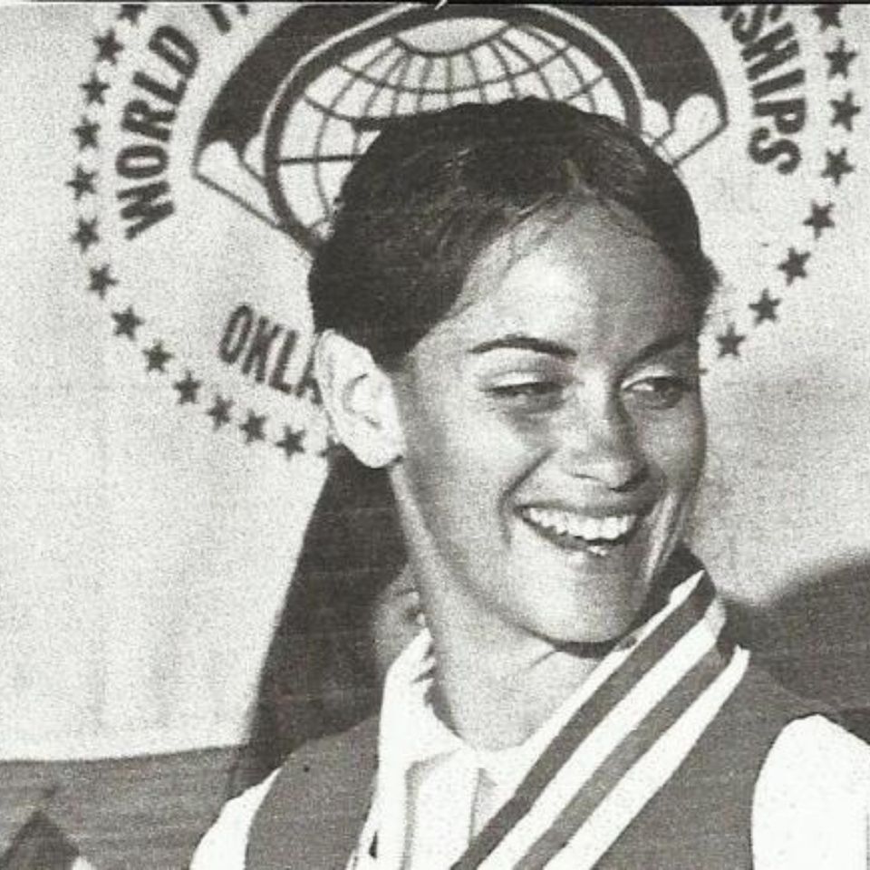 A black and white image of a person smiling, wearing a vest and a medal around their neck. A banner with the text "WORLD..." and "CHAMPIONSHIPS" is partly visible in the background.