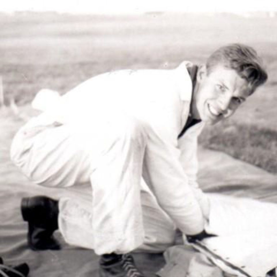 A person wearing a jumpsuit is crouching on the ground outdoors, smiling at the camera. They appear to be preparing or inspecting equipment on a tent or tarp in a grassy area. The image is in black and white.