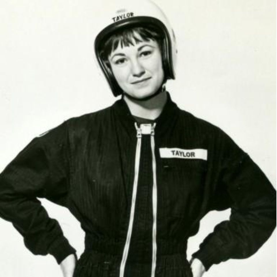 A person wearing a helmet and a racing suit with the name "Taylor" on both the helmet and suit poses confidently with hands on hips. The image is black and white, emphasizing the details of the outfit.