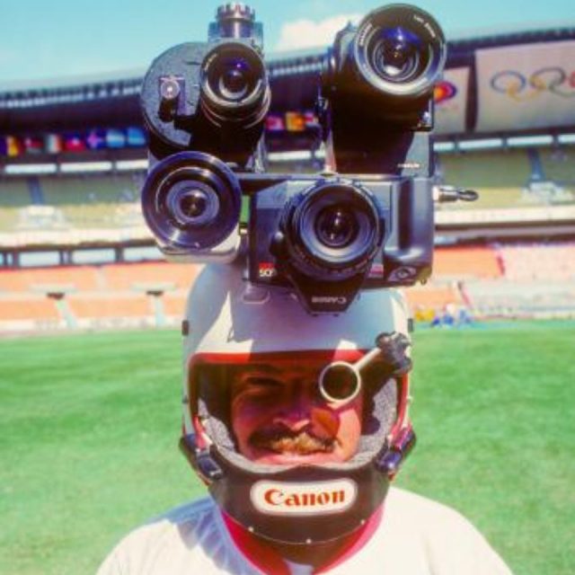 A person in a stadium wears a helmet rigged with multiple cameras on top. The helmet features the Canon logo. The background shows empty stadium seats and an Olympics emblem.