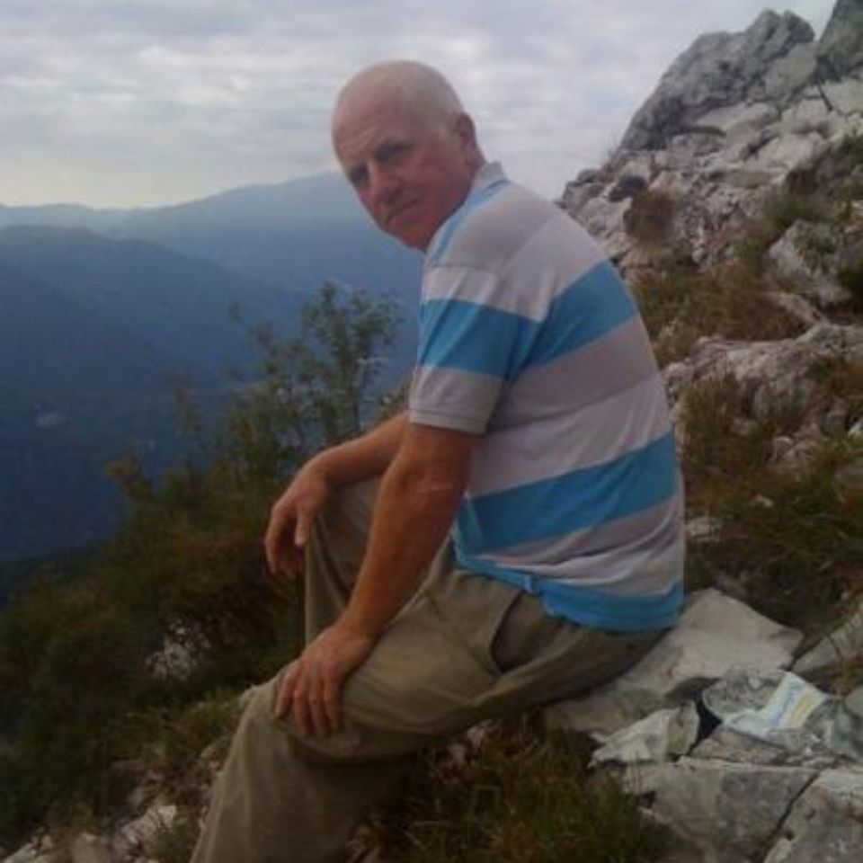 A man wearing a striped shirt and beige pants is sitting on a rocky mountainside. He looks toward the camera with a serene expression. The background features a cloudy sky and distant mountain range.