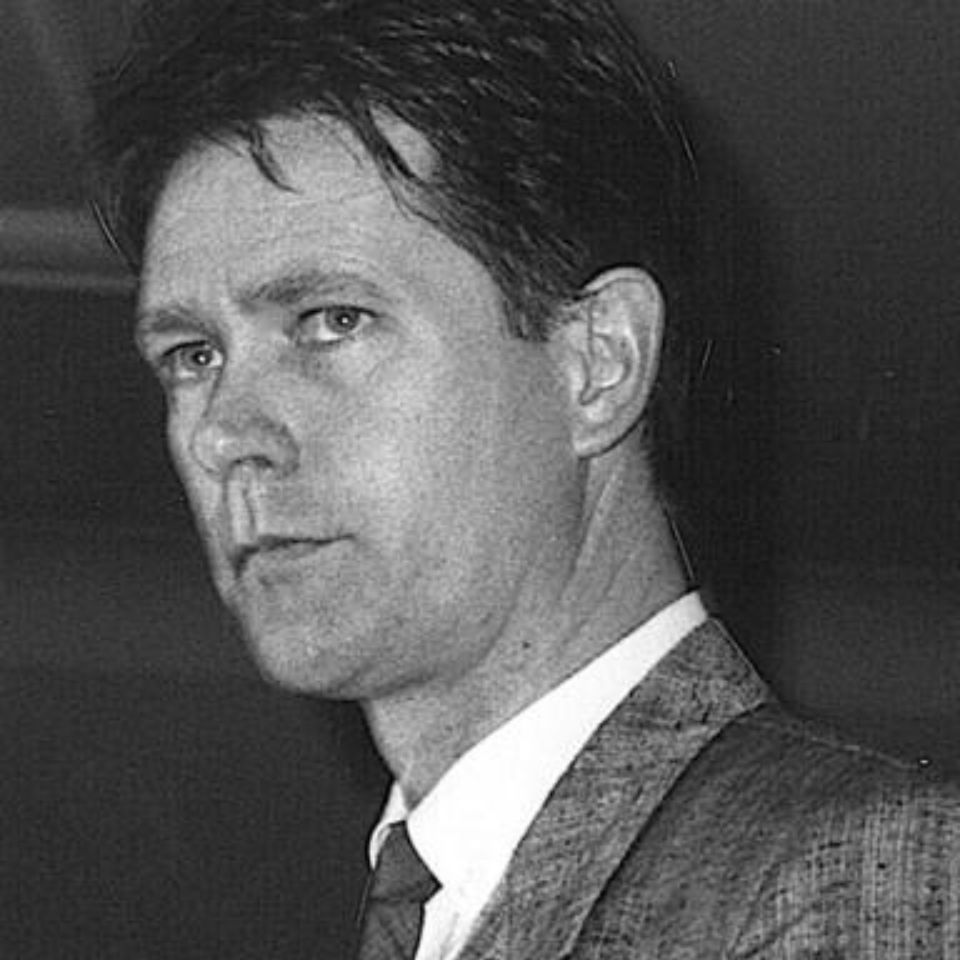 Black and white photo of a man with short hair, wearing a suit and tie, looking slightly to the side with a serious expression.