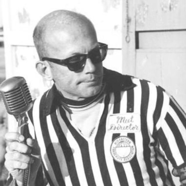 A person wearing sunglasses and a referee uniform holds a microphone. They are looking to the side with a neutral expression. The badge on their shirt is partially visible. The background includes a building or structure. The image is in black and white.