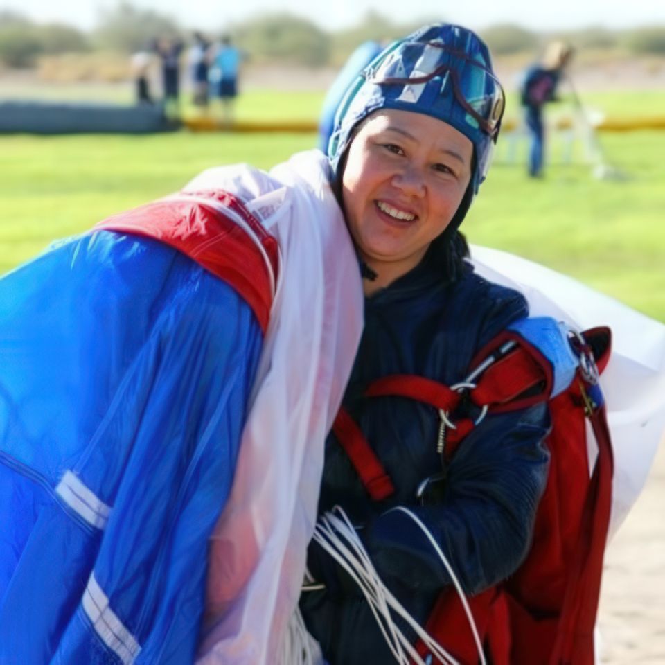 A person smiles while holding a colorful parachute and wearing a helmet and jumpsuit on a sunny day at a skydiving site.