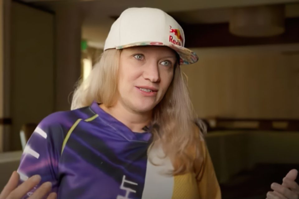 Person wearing a colorful shirt and a white cap with a red logo, gesturing with both hands, indoors.